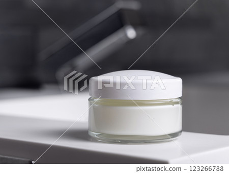 Cream jar with white lid on basin near faucet on basin in dark grey bathroom close up, cosmetic mockup 123266788