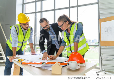 Construction engineer and architect in vest and helmet discussing blueprint with construction manager, standing on construction site. 123266924