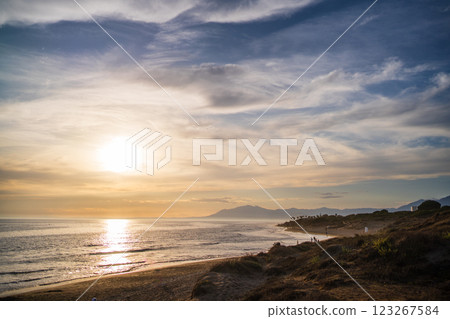 A wide-angle view of a peaceful beach and ocean as the sun sets behind distant hills, casting a warm 123267584