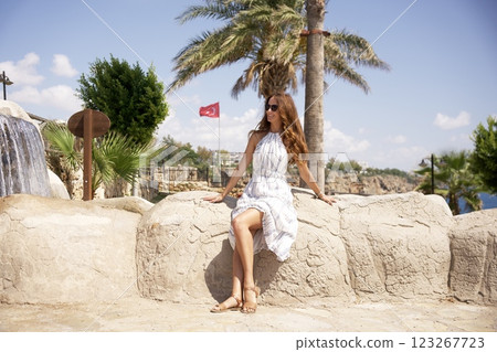 Smiling woman in summer dress sitting by scenic waterfall in Antalya city park 123267723