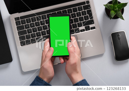 Man holds a smartphone with a green screen above a laptop on a table. Next to a computer mouse, a plant. Place to mockup your application, website Man holds a smartphone with a green screen above a laptop on a table. Next to a computer mouse, a plant. Place to mockup your application, website 123268133