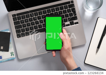Person holds a smartphone with a green screen above a laptop on a table. Nearby a notepad, euro bills, a glass of water. Place to mocap your application, website Person holds a smartphone with a green screen above a laptop on a table. Nearby a notepad, euro bills, a glass of water. Place to mocap your application, website 123268134