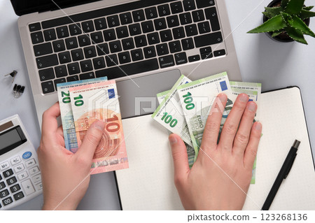 Hands counting euro banknotes over a notebook and notepad on an office desk. Idea passive income, remote work, digital business Hands counting euro banknotes over a notebook and notepad on an office desk. Idea passive income, remote work, digital business 123268136