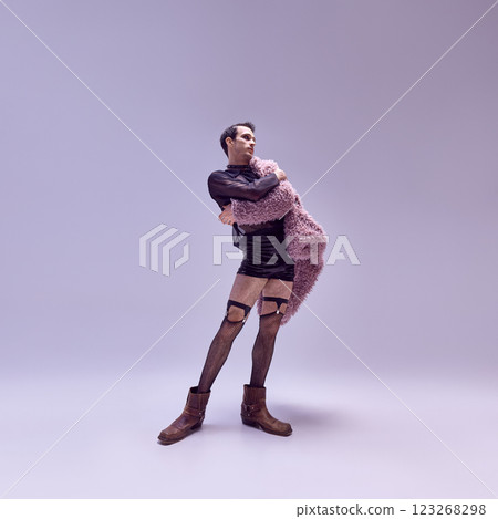 Poised man embracing coat with dramatic stance, wearing bold outfit with high boots against soft-lit backdrop. 123268298