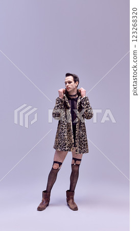Artistic young man standing in leopard coat and fishnet tights with fierce expression and confident stance against white studio background. Artistic young man standing in leopard coat and fishnet tights with fierce expression and confident stance against white studio background. 123268320