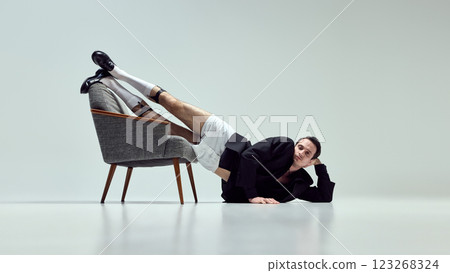 Expressive young man, dressed stylish outfit, leaning off chair with relaxed yet unusual posture against white studio background. 123268324