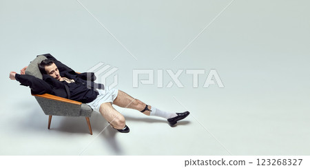 Fashion-forward young man stretching in chair with relaxed posture and vintage aesthetic against white studio background. 123268327