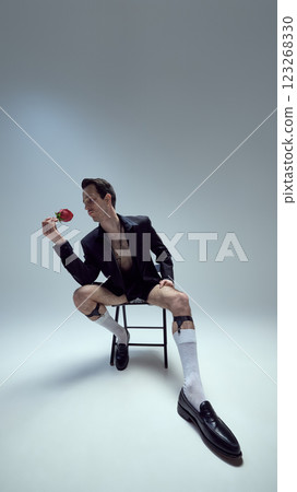 Confident young man posing, sitting on stool admiring rose with stylish pose and sharp fashion against white studio background. Confident young man posing, sitting on stool admiring rose with stylish pose and sharp fashion against white studio background. 123268330