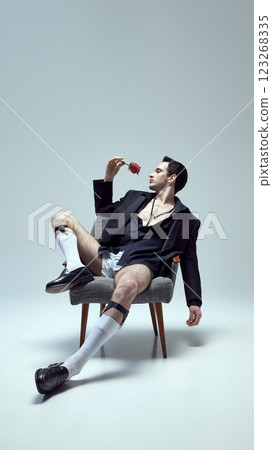 Stylish, handsome man posing, reclining on chair holding rose with intense gaze and relaxed posture against white studio background. 123268335