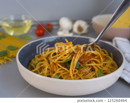 Lifting balanesi pasta with tomato sauce and basil from a modern bowl, surrounded by blurred ingredients like uncooked spaghetti and cherry tomatoes 123268464