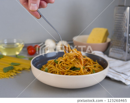 Professional chef lifting balanesi pasta with fork, surrounded by kitchen ingredients: tomatoes, mushrooms, olive oil, cheese, basil, raw spaghetti on wooden surface 123268465