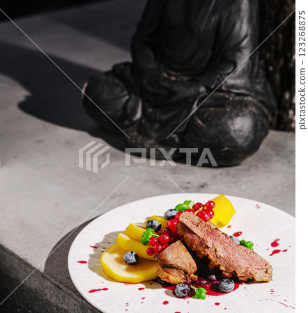 Gourmet dish featuring fruits and meat next to a serene statue 123268875