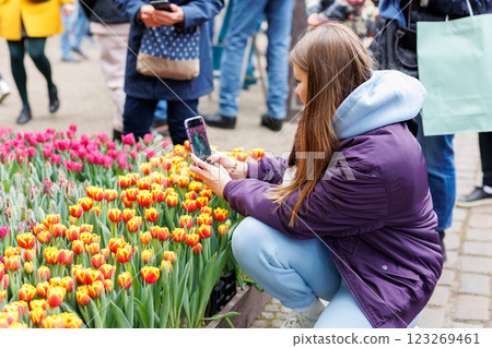 Young woman with smartphone leans over bright tulips in bustling outdoor event. She captures colorful blossoms while public gathers behind, enjoying vibrant spring display and seasonal beauty 123269461
