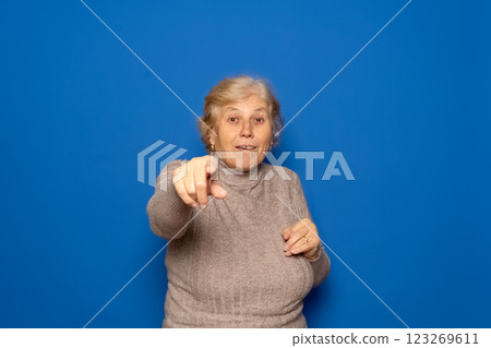 Caucasian woman in her 70s wearing a dark beige sweater pointing cheerfully at the camera, isolated over blue studio . 123269611