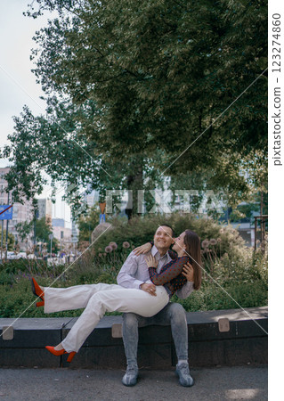 Happy couple in love enjoying a playful moment in the city park. A young man sitting on a bench holding his girlfriend in his arms as they smile at each other. Urban nature, romance, and joy. 123274860