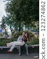 Happy couple in love enjoying a playful moment in the city park. A young man sitting on a bench holding his girlfriend in his arms as they smile at each other. Urban nature, romance, and joy. 123274862