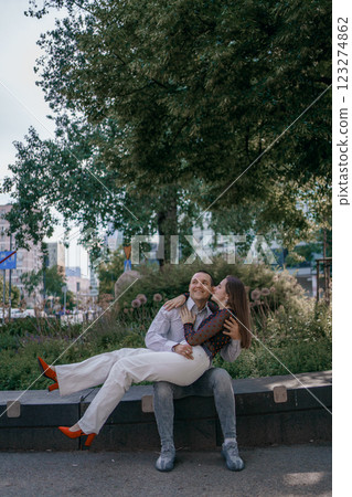 Happy couple in love enjoying a playful moment in the city park. A young man sitting on a bench holding his girlfriend in his arms as they smile at each other. Urban nature, romance, and joy. 123274862