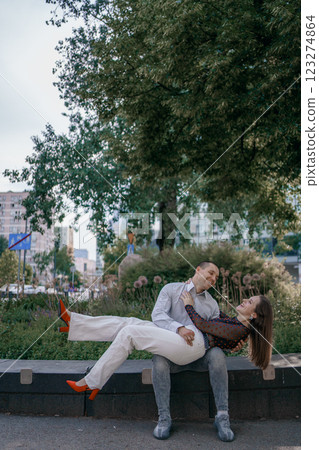 Happy couple in love enjoying a playful moment in the city park. A young man sitting on a bench holding his girlfriend in his arms as they smile at each other. Urban nature, romance, and joy. 123274864