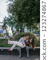 Happy couple in love enjoying a playful moment in the city park. A young man sitting on a bench holding his girlfriend in his arms as they smile at each other. Urban nature, romance, and joy. 123274867