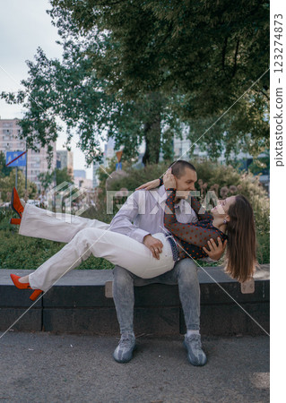 Happy couple in love enjoying a playful moment in the city park. A young man sitting on a bench holding his girlfriend in his arms as they smile at each other. Urban nature, romance, and joy. Happy couple in love enjoying a playful moment in the city park. A young man sitting on a bench holding his girlfriend in his arms as they smile at each other. Urban nature, romance, and joy. 123274873