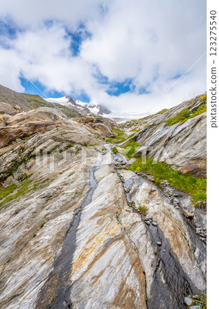 A stunning view of the Schlaten Glacier area in Austria, featuring rugged, layered rocky terrain illuminated by soft sunlight under a partly cloudy sky. The landscape showcases the beauty of Alps 123275540