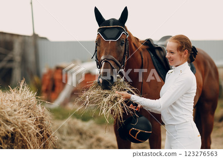 Woman rider jockey in white uniform feeds horse at stable 123276563