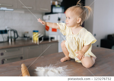 Playful toddler girl sitting amid scattered flour, reaching hands across messy kitchen table, exploring baking ingredients with curious expression. Concept of simple pleasures of childhood. Playful toddler girl sitting amid scattered flour, reaching hands across messy kitchen table, exploring baking ingredients with curious expression. Concept of simple pleasures of childhood. 123276732