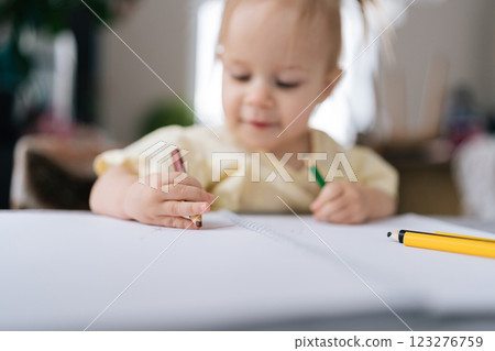Selective focus shot of cute toddler girl happily drawing with colorful pencils at home sitting at desk by window, developing creativity, imagination, fine motor skills in playful and engaging way. Selective focus shot of cute toddler girl happily drawing with colorful pencils at home sitting at desk by window, developing creativity, imagination, fine motor skills in playful and engaging way. 123276759
