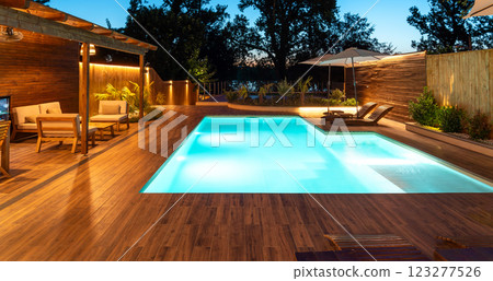Luxurious backyard pool with a wooden deck and cozy lounge area. Evening retreat illuminated with warm, inviting lighting 123277526