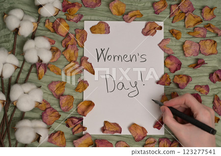 Woman's hand with a pen writing "Women's Day!" on white paper. Green cloth background with dry rose petals and cotton inflorescence plant. Greeting, invitation card. Top view Woman's hand with a pen writing "Women's Day!" on white paper. Green cloth background with dry rose petals and cotton inflorescence plant. Greeting, invitation card. Top view 123277541