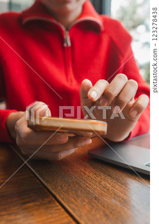 A person in a red sweater uses a smartphone at a wooden table, with a laptop in the background. 123278438