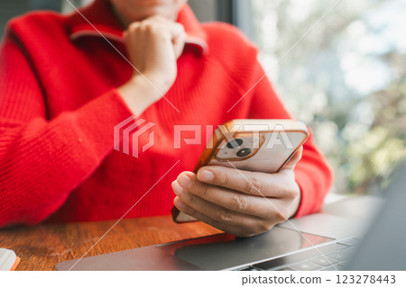 Close-up of a woman in a red sweater using a smartphone at a wooden table with a laptop, creating a cozy indoor atmosphere. Close-up of a woman in a red sweater using a smartphone at a wooden table with a laptop, creating a cozy indoor atmosphere. 123278443
