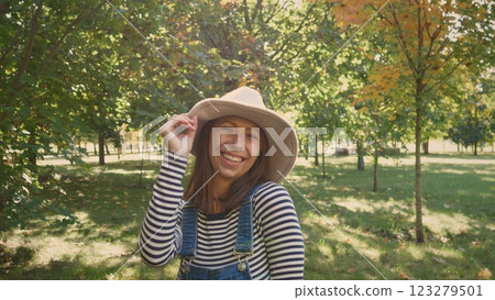 A joyful woman donning a stylish hat delights in the beautiful sunny day spent in a vibrant lush green park 123279501