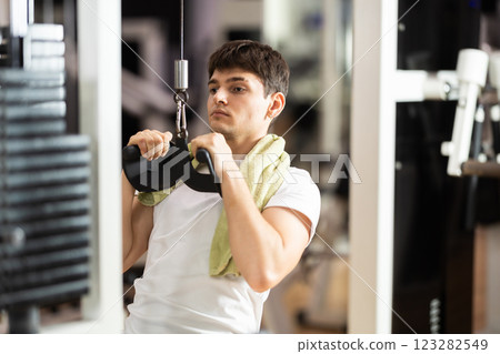 Guy in gym performs exercises for arm muscles using cable crossover machine and pull weighted rope Guy in gym performs exercises for arm muscles using cable crossover machine and pull weighted rope 123282549