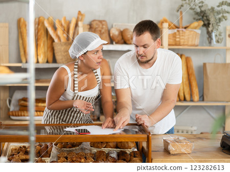 In bakery, male visitor and female salesperson work with paper documents and count using phone 123282613