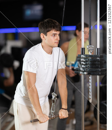 Guy in gym performs exercises for arm muscles using cable crossover machine and pull weighted rope 123282867