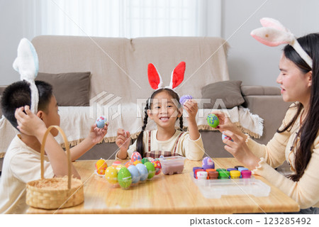 A mother and kids with bunny ears happily paint Easter eggs at a table near a festive basket A mother and kids with bunny ears happily paint Easter eggs at a table near a festive basket 123285492