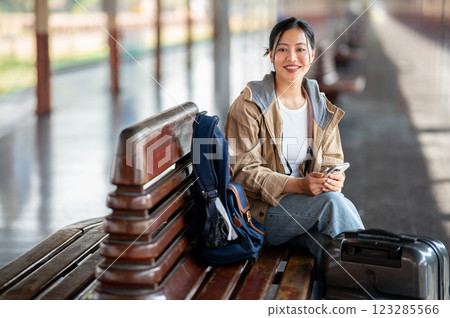 An Asian woman is smiling with the phone in hand while sitting on the wooden train station bench. An Asian woman is smiling with the phone in hand while sitting on the wooden train station bench. 123285566