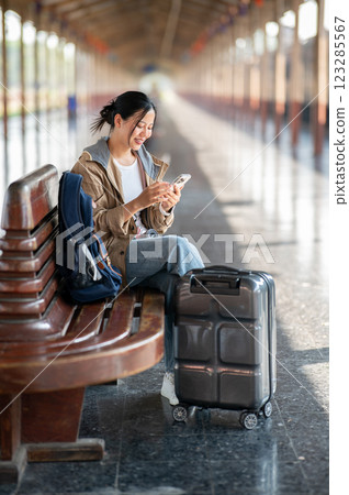 Asian woman is looking at the phone while sitting on wooden train station bench with bag and luggage 123285567