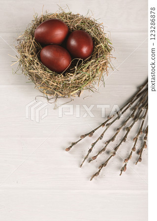 Red colored eggs in birds nest and pussy willow on wooden white background, top view, vertical image. Atmospheric Easter concept. 123286878
