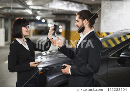 Caucasian businesswoman selling car to man handing over keys in underground parking lot. Professional attire and successful transaction in automotive setting. Caucasian businesswoman selling car to man handing over keys in underground parking lot. Professional attire and successful transaction in automotive setting. 123288304