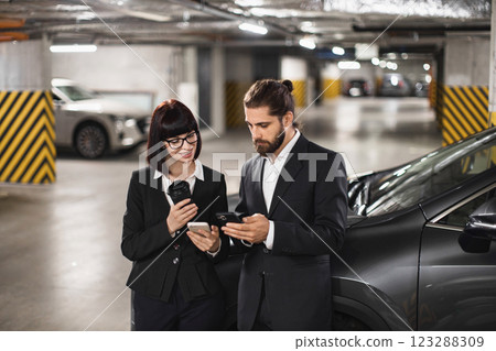 Caucasian male and female professionals using smartphones in urban parking garage. Business attire, modern technology, focused engagement. Teamwork, networking, communication in work environment. 123288309