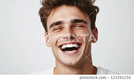 Young man with contagious joy, eyes closed, beaming smile showcasing healthy teeth, brown hair, carefree expression against a bright, plain background. 123291250