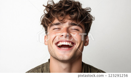 Joyful expression: A young man with curly hair laughs heartily, conveying genuine happiness and exuberance against a plain backdrop. 123291256