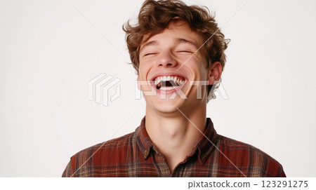 A joyful young man with curly hair and a plaid shirt is laughing heartily against a plain white backdrop, radiating pure happiness. 123291275