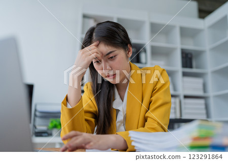 Stressed businesswoman in yellow blazer overwhelmed by work at a desk with papers and laptop in a modern office setting 123291864