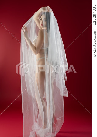 Beautiful bride in delicate lingerie with flowing veil against red backdrop 123294339
