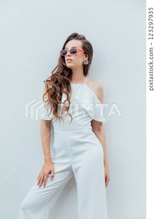 Beautiful brunette woman in white suit posing by the wall with plants Beautiful brunette woman in white suit posing by the wall with plants 123297935