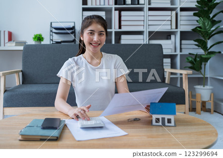 Young woman analyzing financial documents at home with calculator and model house on table in modern living room 123299694
