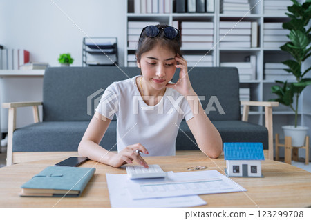 Young woman analyzing financial documents at home with calculator and model house on table in modern living room Young woman analyzing financial documents at home with calculator and model house on table in modern living room 123299708